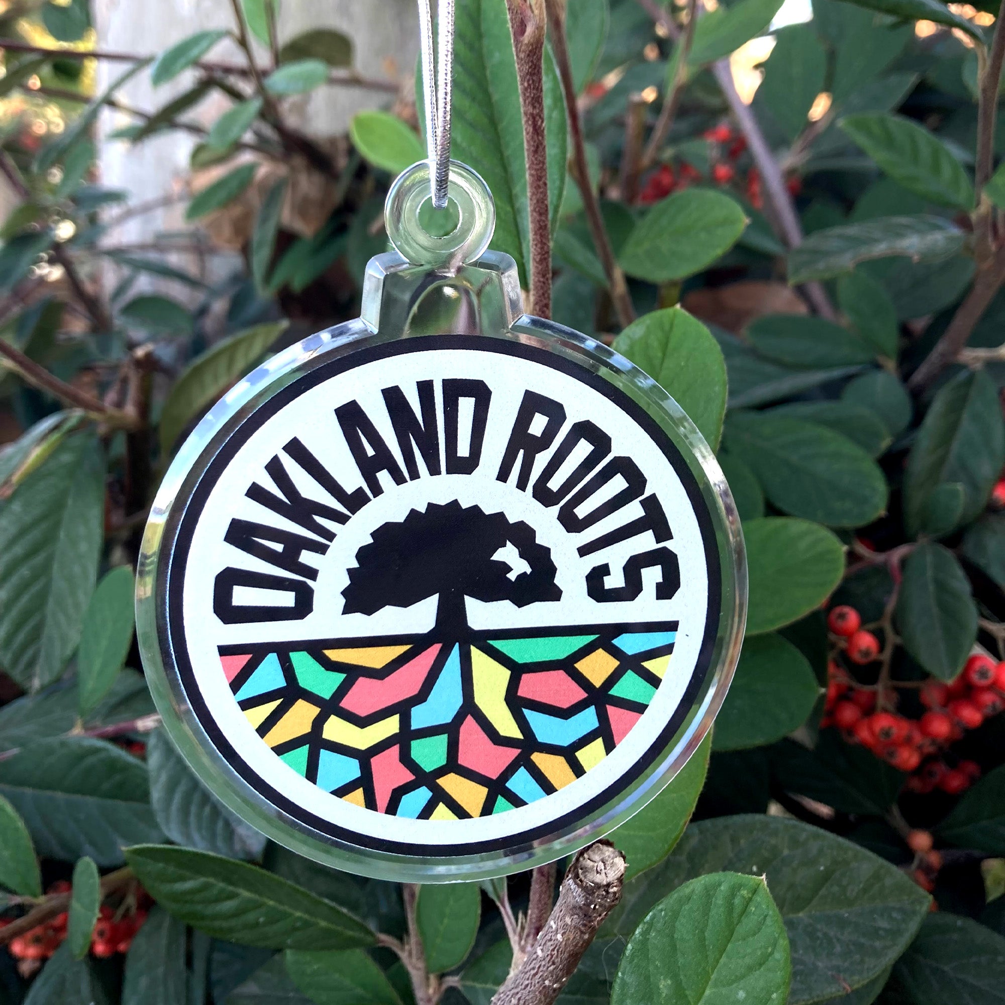 Acrylic Oakland Roots SC logo crest on a holiday ornament with silver string hanging on a holly bush.