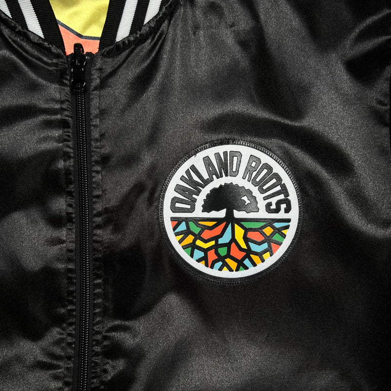 Full-color Roots SC embroidered logo patch on the chest of a black satin Mitchell & Ness jacket.