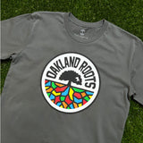 Three-quarter view of a charcoal grey t-shirt with full-color, round Oakland Roots logo on the chess lying on the grass.