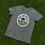 Charcoal grey youth t-shirt with full-color Roots SC circle logo on the chest laying on green grass.