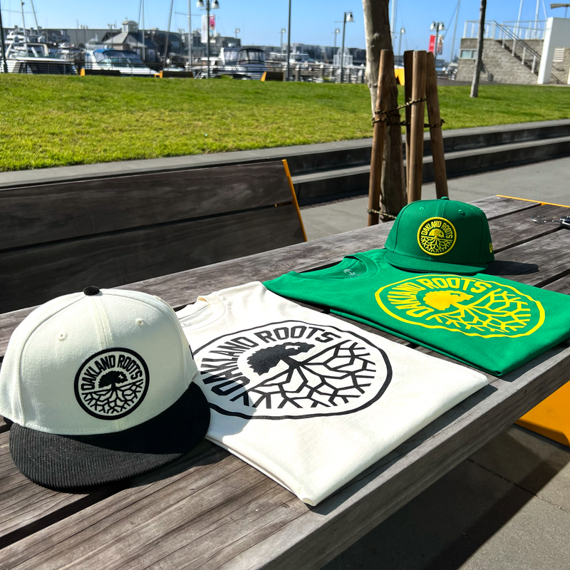 Kelly green and Ecru t-shirts with large Oakland Roots SC crests and two New Era caps in Kelly green and Chrome white on an outdoor picnic table.
