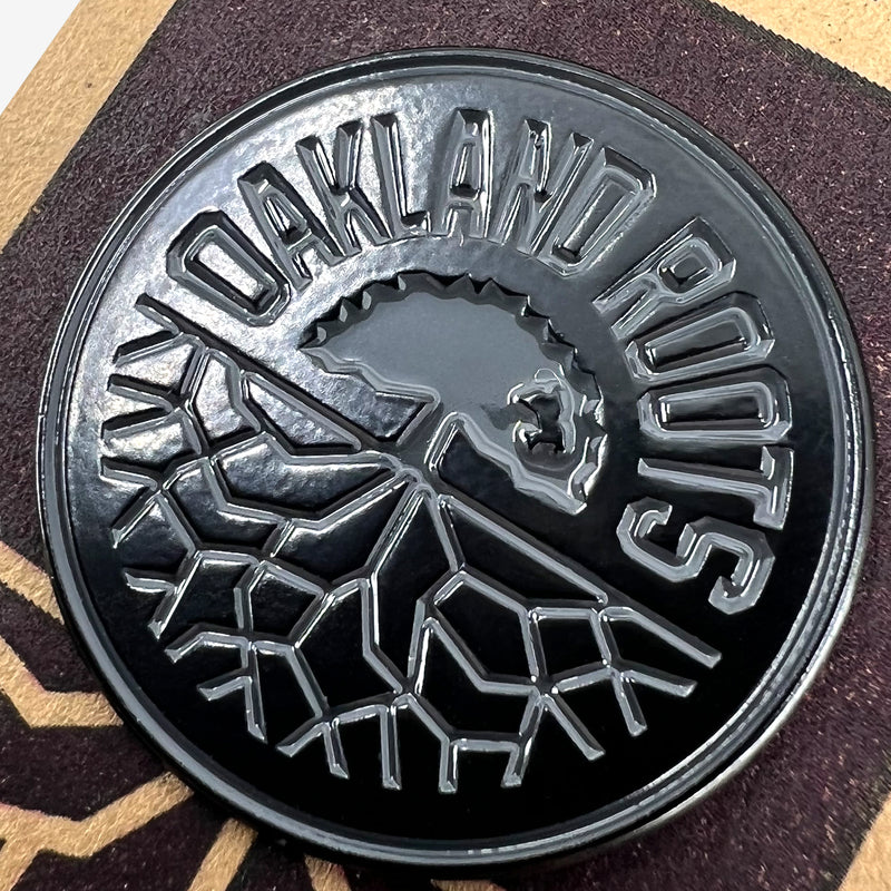 Close-up of black enamel lapel pin with round black Oakland Roots logo on brown paper retail packaging. 