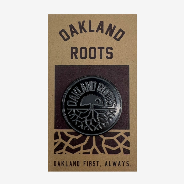 Black enamel lapel pin with round black Oakland Roots logo on brown paper retail packaging with words Oakland First, Always. 