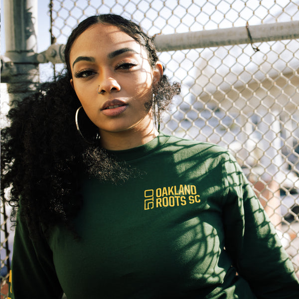 Women leaning against an outdoor fence wearing a long-sleeved green t-shirt with 510 Oakland Roots SC on the chest.