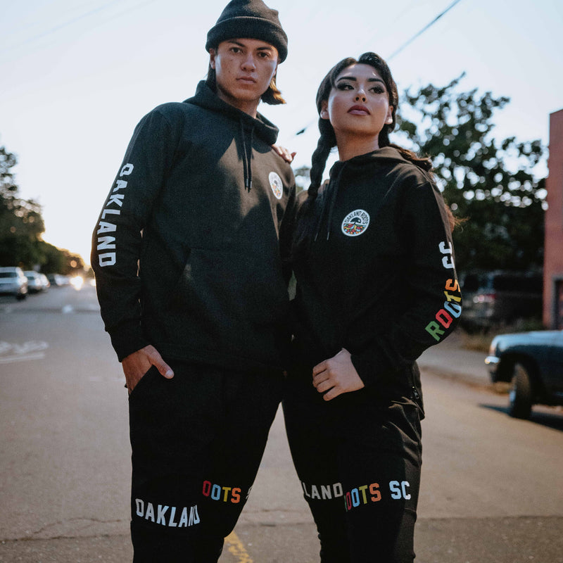 Man and woman standing on street wearing black Oakland Roots SC hoodie and jogger sets.