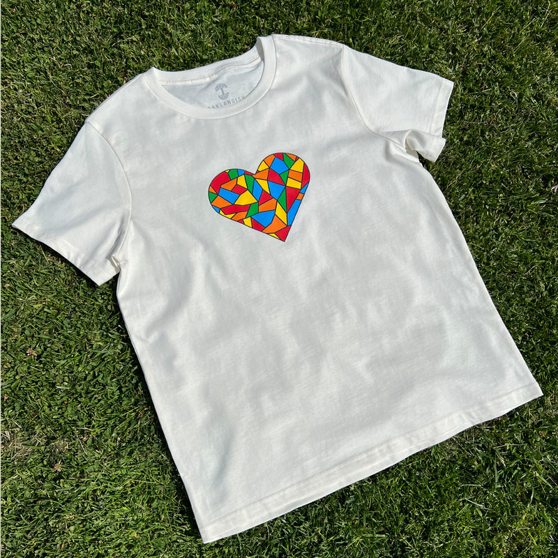 Natural cotton t-shirt with full-color Roots mosaic logo colors in a heart shape on chest laying on grass.