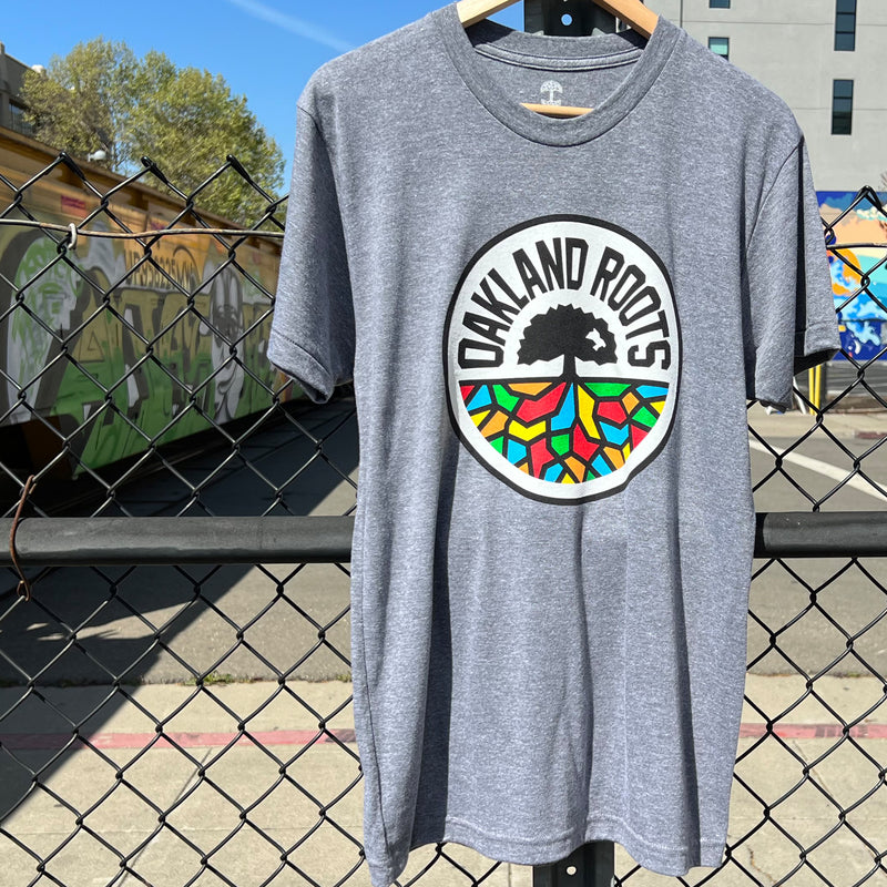 Grey t-shirt with full-color, round Oakland Roots logo on the chest hanging on a chain fence in an urban area with graffiti.