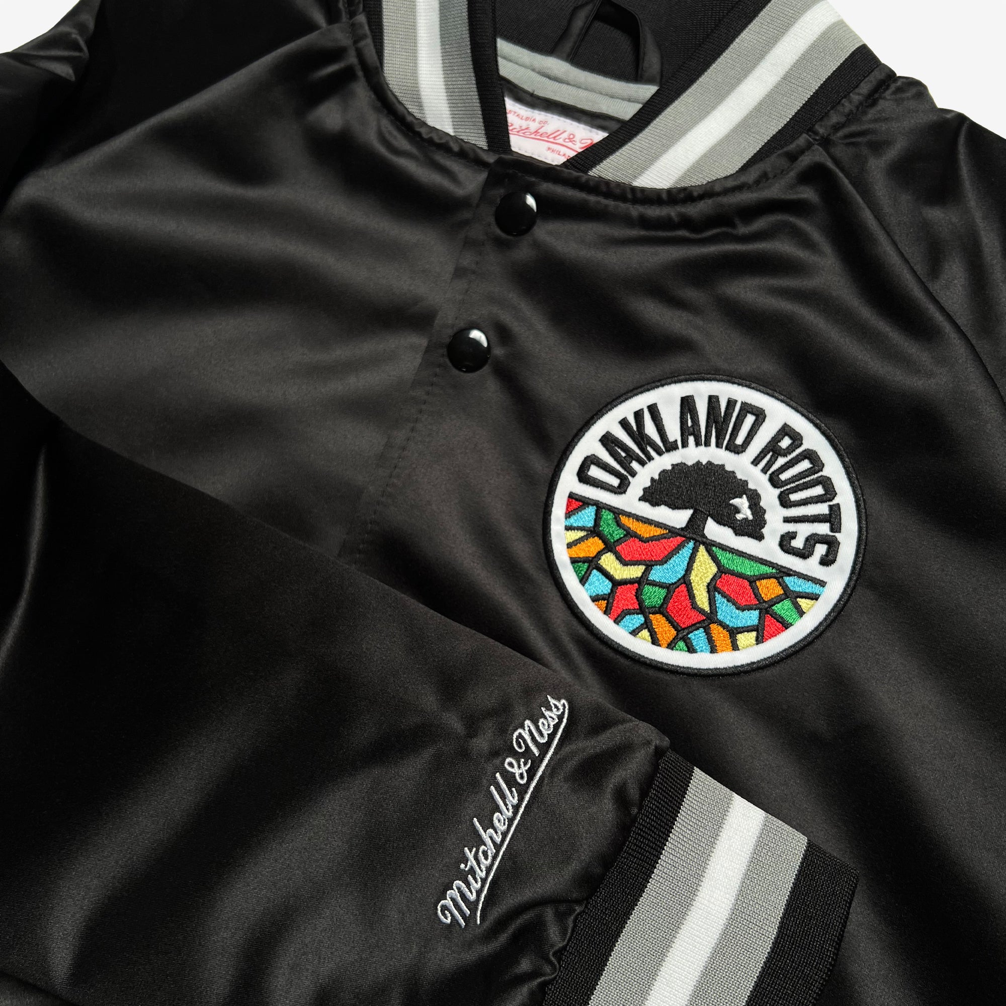 The Mitchell & Ness Oakland Roots SC Lightweight Satin Jacket features a sleek black satin finish, highlighted by a round embroidered "Oakland Roots" patch with a vibrant pattern. The jacket has gray and white striped collar, cuffs, and waistband, while the sleeve showcases "Mitchell & Ness" embroidery in gray. This sports club jacket is designed with front snaps for easy fastening.