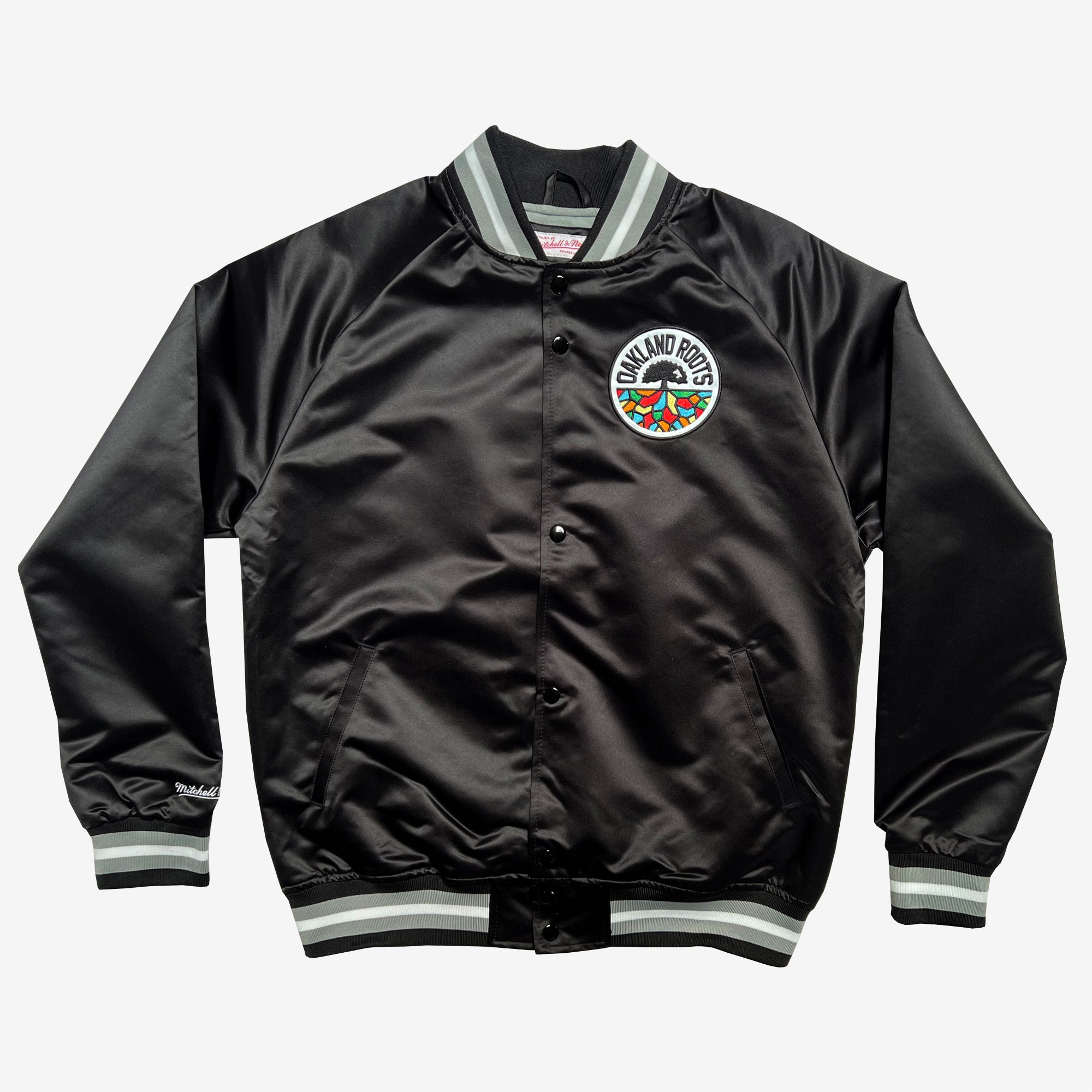 The Mitchell & Ness Oakland Roots SC Lightweight Satin Jacket is designed with black satin fabric, ribbed cuffs, and waistband, accented by green, white, and grey stripes. It features a snap-button closure and an embroidered circular patch on the left chest that showcases multi-colored stylized text reading "OAKLAND ROOTS" alongside a tree design. This jacket is perfect for representing your favorite sports club.