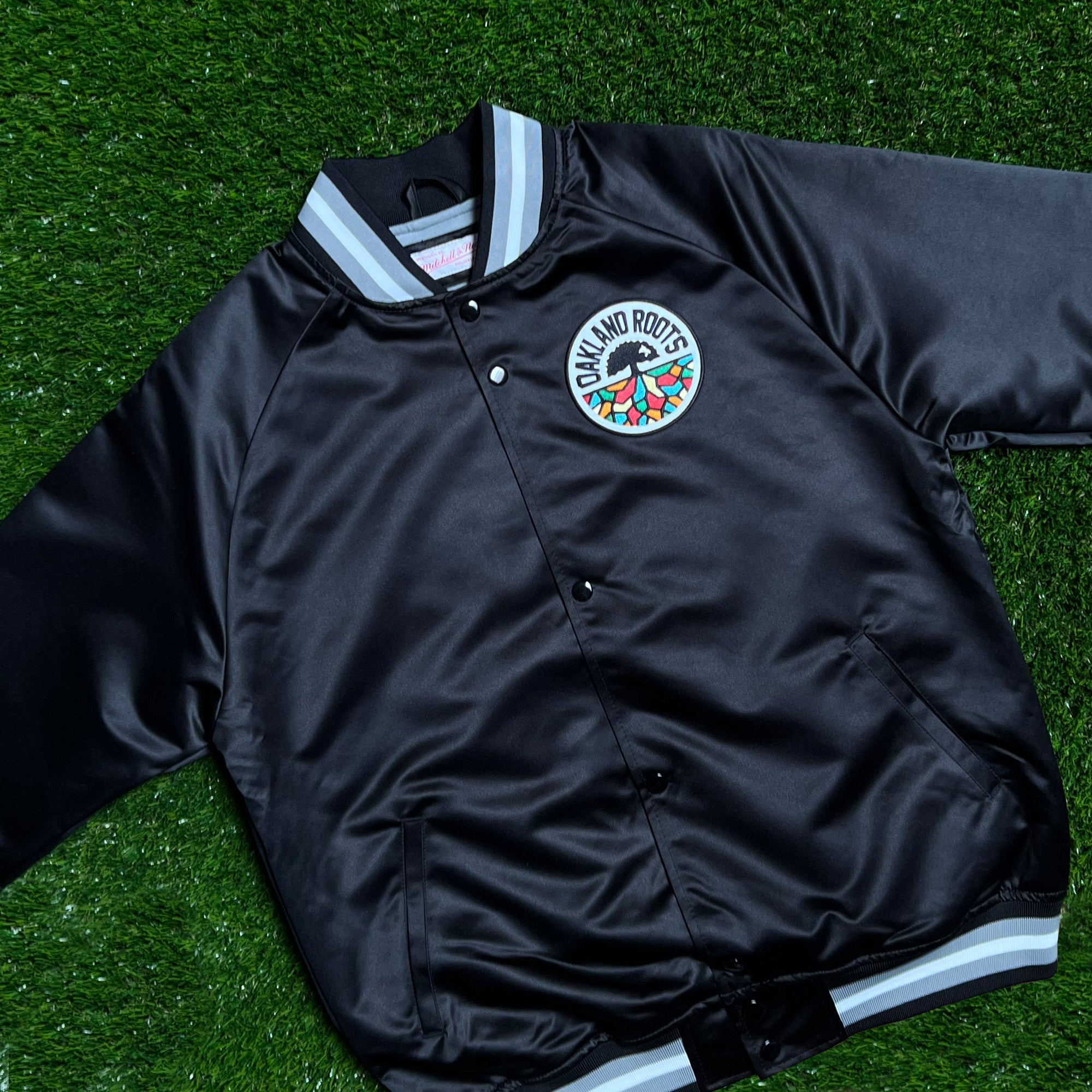 The Mitchell & Ness Oakland Roots SC Lightweight Satin Jacket, featuring a black satin design with gray and white striped ribbed collar, cuffs, and hem. This lightweight jacket includes a colorful circular "Oakland Roots" logo patch on the left chest, showcasing a tree with diverse colored roots. Perfect for any sports club fan, the jacket is laid flat on vibrant green grass.