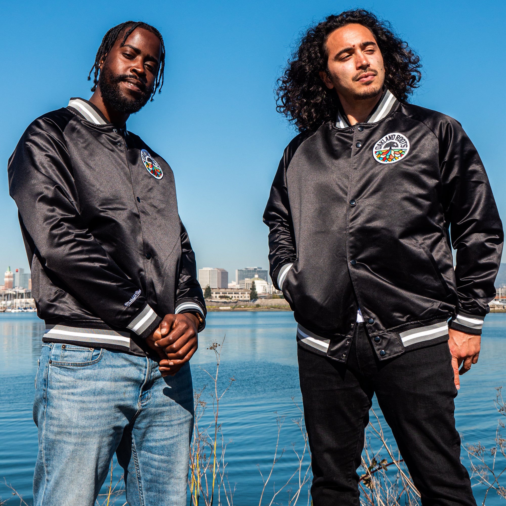 Two men stand in front of a body of water with a cityscape in the background, both wearing the Mitchell & Ness Oakland Roots SC Lightweight Satin Jacket, identifiable by its striped cuffs. The man on the left, sporting a beard and light blue jeans, has an Oakland Roots patch on his jacket. The man on the right with curly hair wears dark jeans. Both have serious expressions.