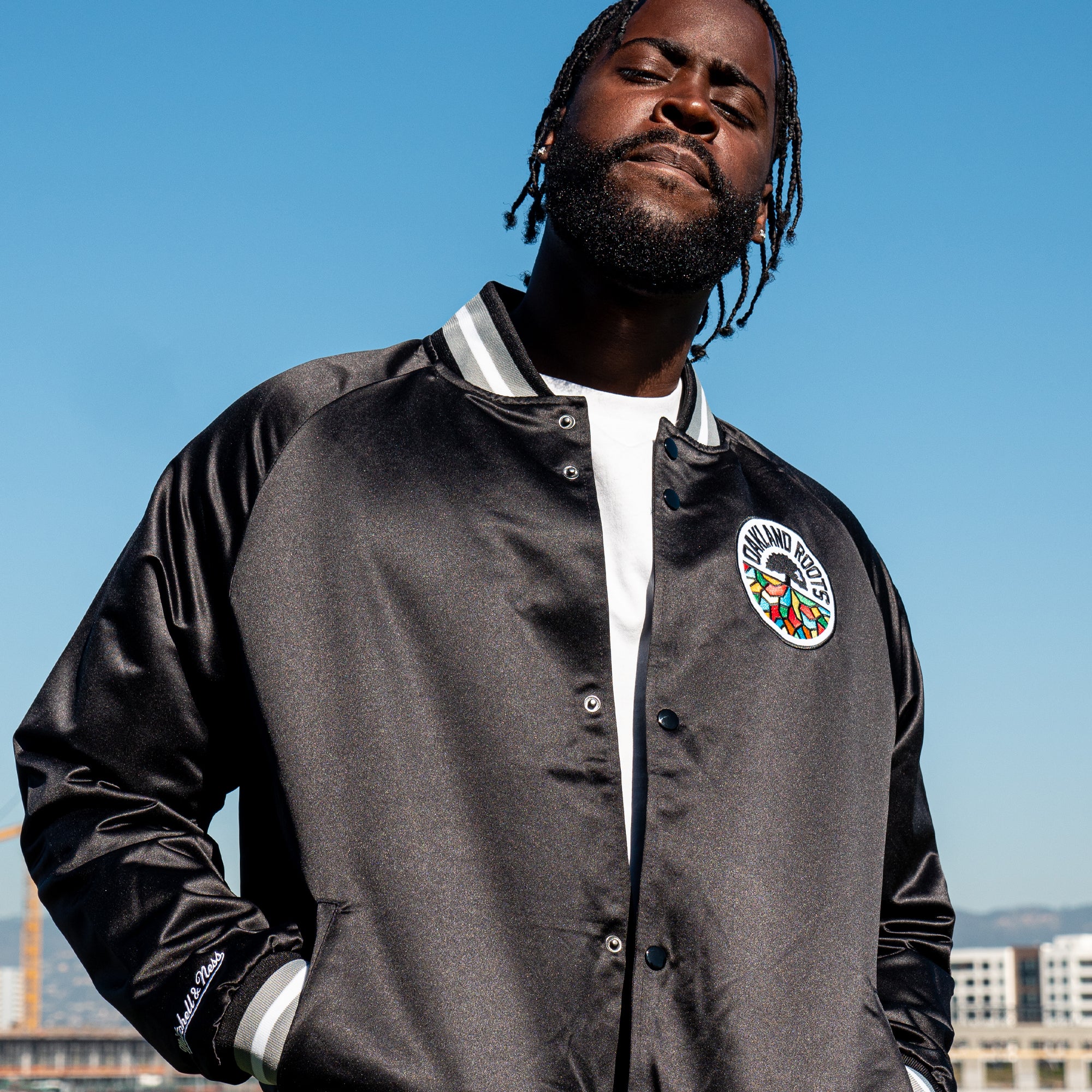 A person wearing a Mitchell & Ness Oakland Roots SC Lightweight Satin Jacket stands confidently against a clear blue sky. The black varsity jacket, from the Oakland Roots SC brand, features white stripes on the collar and cuffs and an Oakland Roots patch on the chest. They are looking slightly upward with a calm expression, while city buildings can be seen in the background.