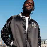 Man in a black satin jacket with striped collar, sleeve cuffs, buttons, and round Roots SC logo with hands in front pockets.