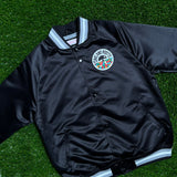 A black satin jacket with a striped collar and round Roots SC logo patch lying on the grass.