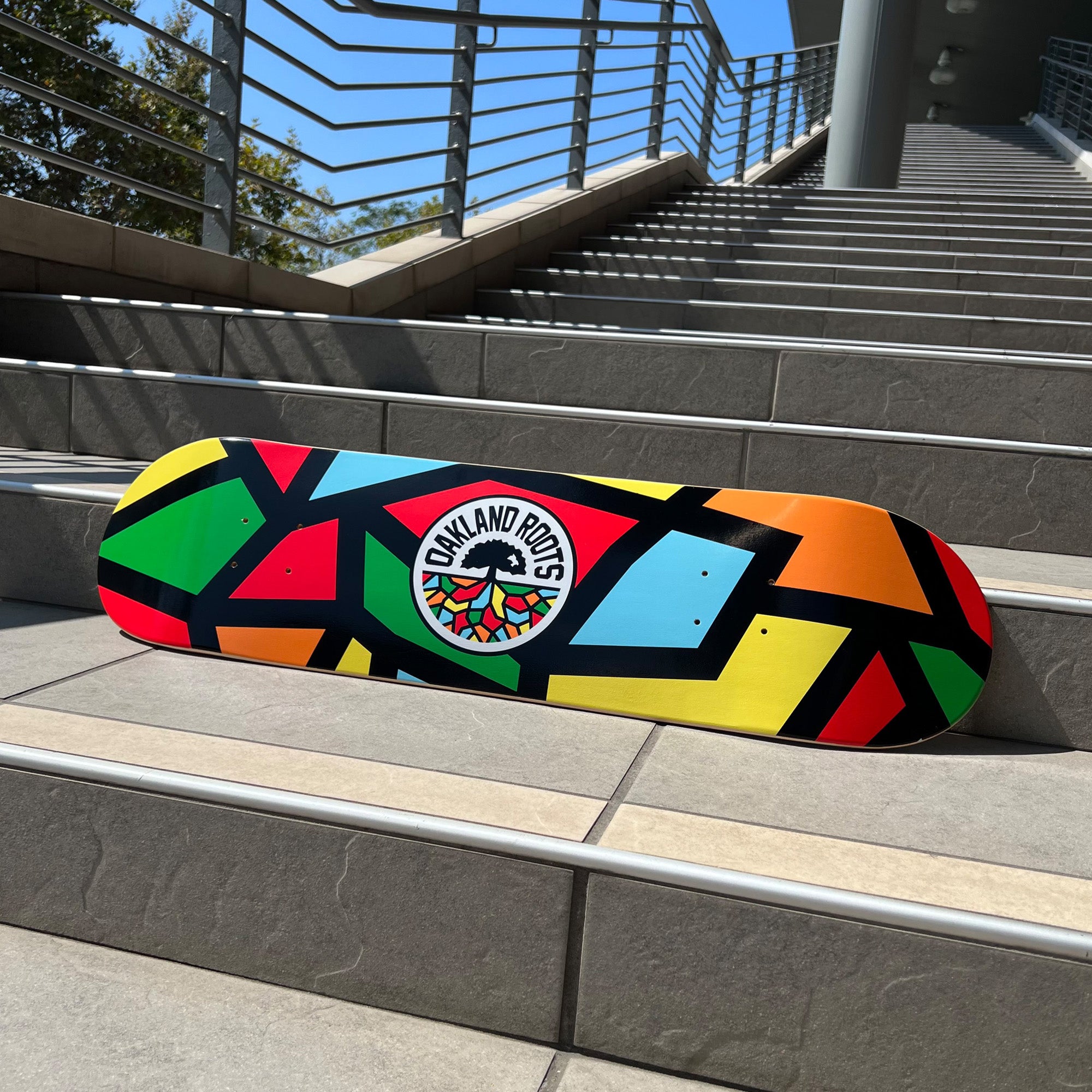 Skateboard deck with full-color Roots Soccer Club mosaic colors and round logo mark laying outside on stairs.