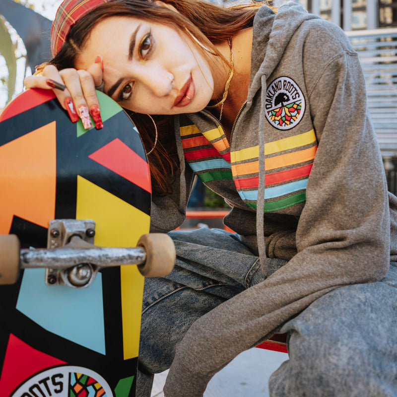 Woman outside with skateboard wearing gunmetal heather full zip hoodie with colored stripes and full-circle Roots SC mosaic logo on the chest.