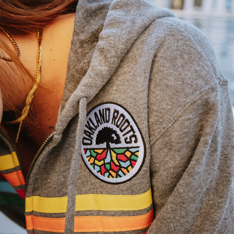 Detailed close up of women's chest outside wearing gunmetal heather full zip hoodie with colored stripes and full-circle Roots SC mosaic logo.