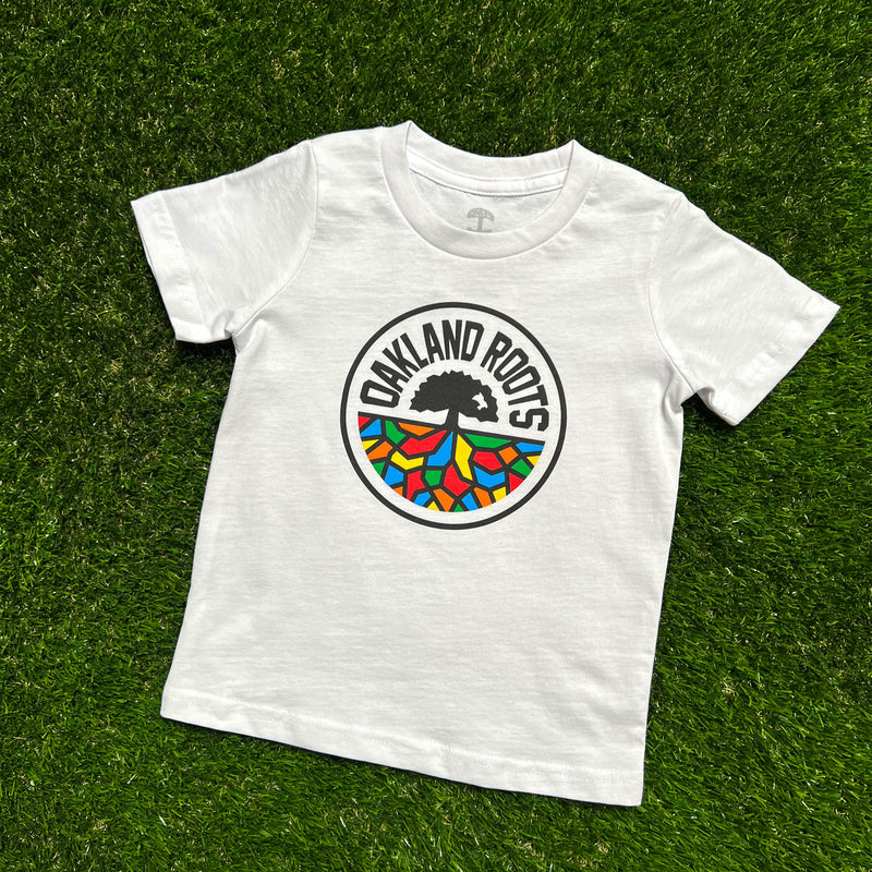 White toddler-sized t-shirt with a full-color Roots SC logo on the chest laying on grass.