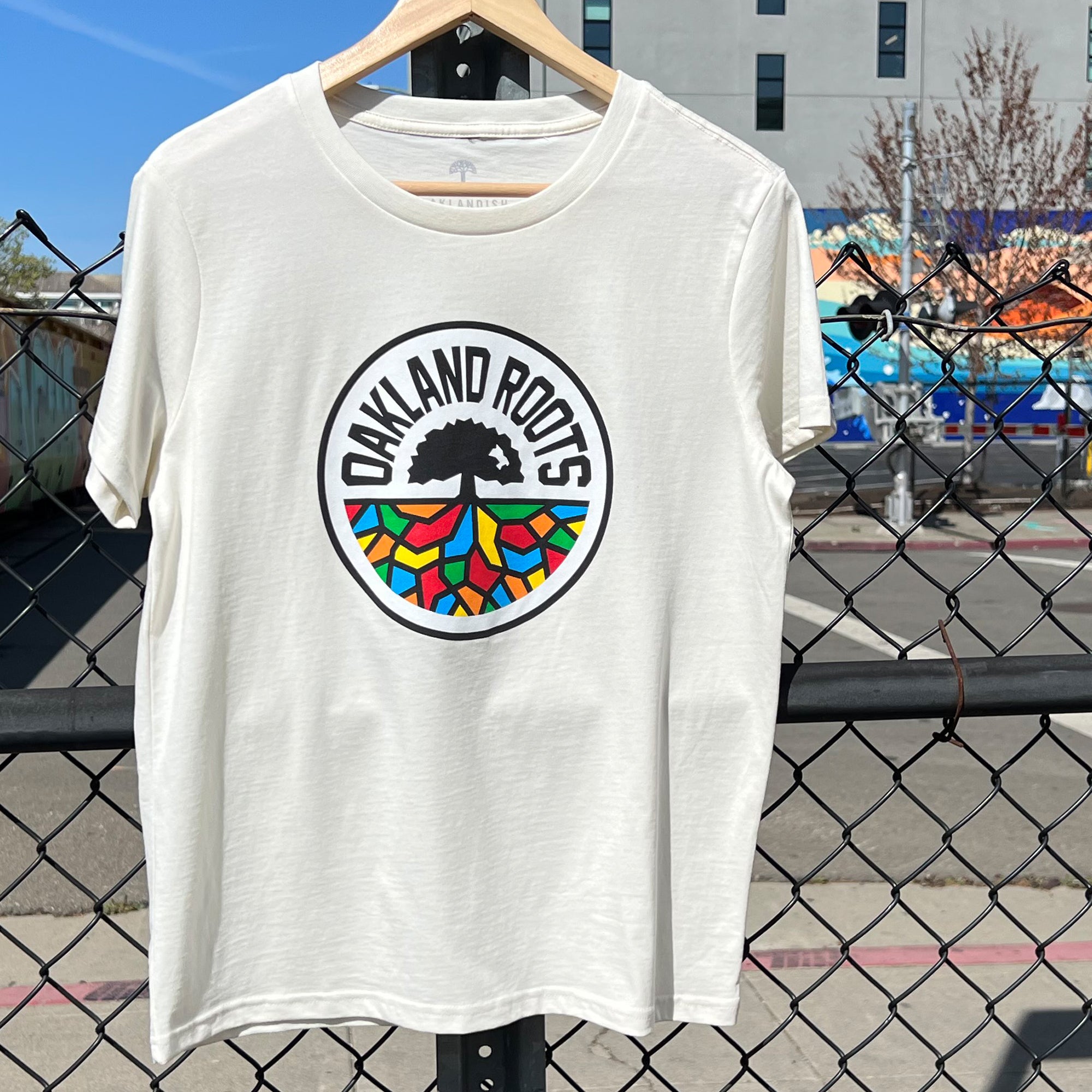 A natural colored women’s t-shirt with a full-color Roots SC logo on the chest hanging on an urban chain link fence. 