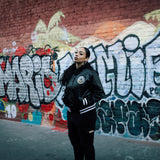 Woman standing in front of a graffiti wall wearing a Mitchell & Ness Black satin zip-up reversible jacket with Oakland Roots crest on the chest.