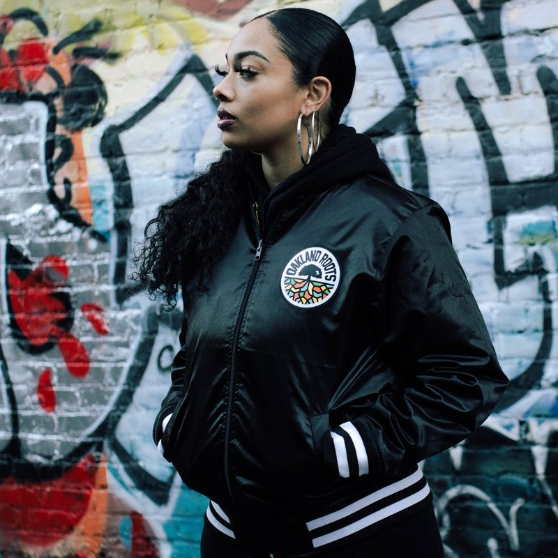 Woman standing outside wearing Mitchell & Ness Black satin zip-up reversible jacket with Oakland Roots crest on the chest and striped ribbing on the collar, cuff, and waistband.