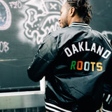 A man standing outside, back to the camera, wearing Mitchell & Ness black satin jacket with a large centered Oakland Roots wordmark.