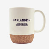 OAKLANDISH CERAMIC HOME GOODS PROCURED FOR THE TOWN on a cream colored ceramic mug with cork bottom outdoors on a picnic table.