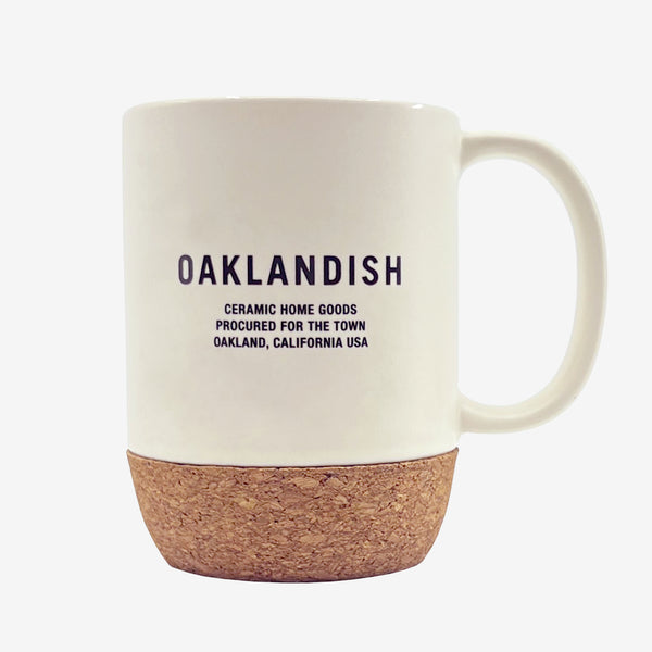 OAKLANDISH CERAMIC HOME GOODS PROCURED FOR THE TOWN on a cream colored ceramic mug with cork bottom outdoors on a picnic table.