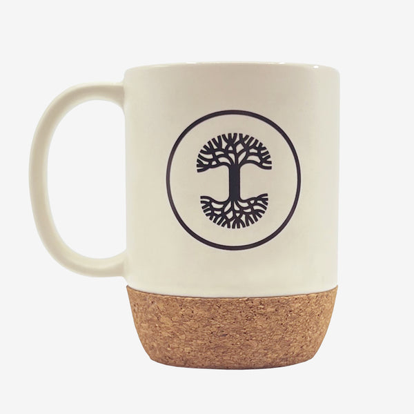 Cream colored rustic ceramic mug with black Oaklandish tree logo in a circle and cork bottom outdoors on a picnic table.