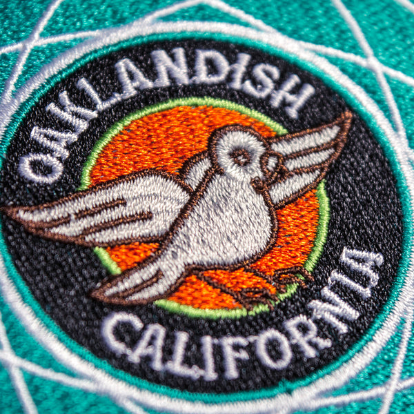 Round embroidered iron-on patch with turquoise blue outer rim, white pigeon in middle orange and grey circles, and “Oaklandish California” wordmark. 