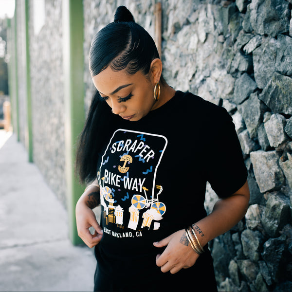 Female model wearing black t-shirt with blue, white & yellow Scraper Bike Way graphic & East Oakland wordmark