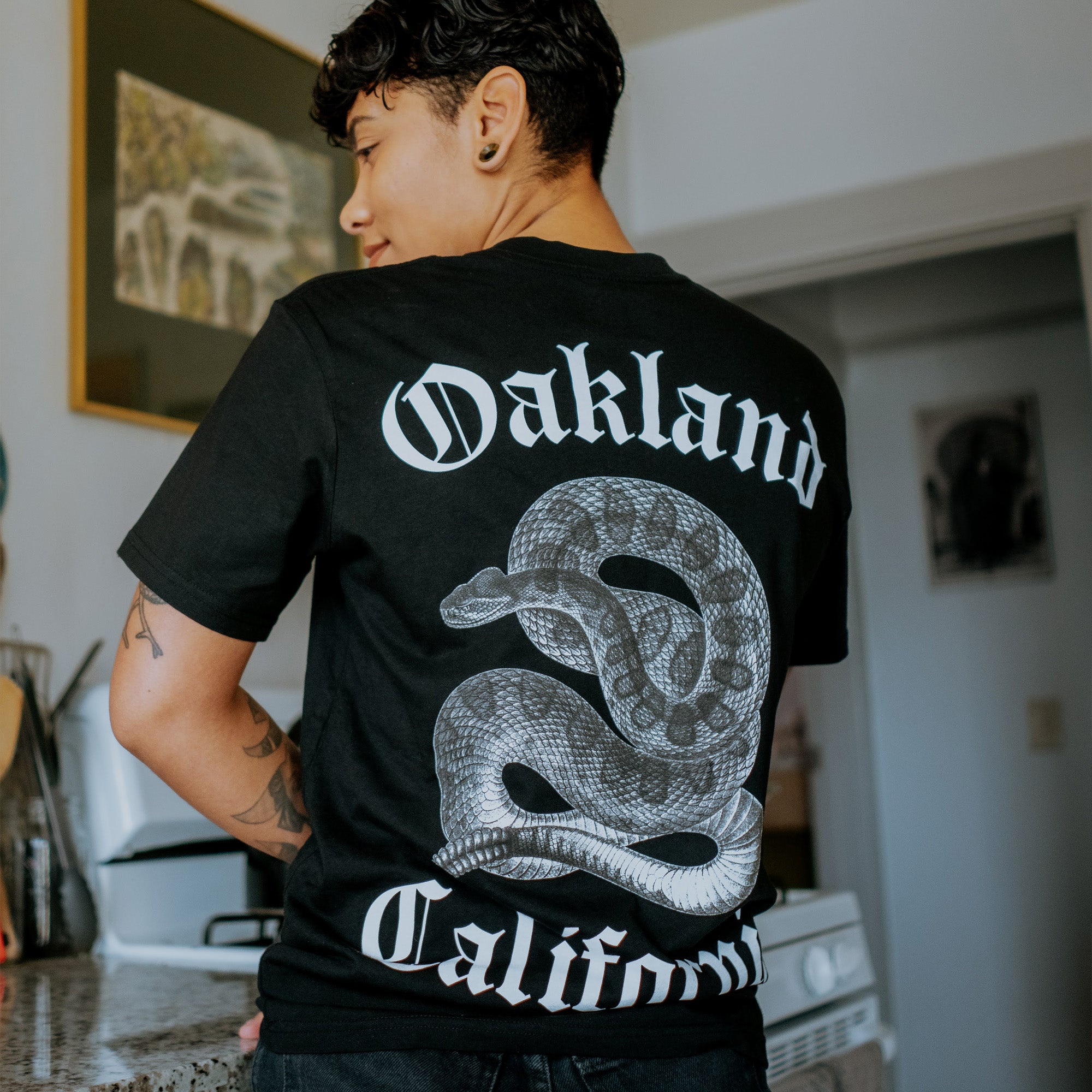 A person with short, curly hair is standing sideways in a kitchen, wearing an Oaklandish Sistrurus Tee. The classic fit T-shirt, made from 100% cotton, features the text "Oakland California" in white along with a large venomous snake graphic coiled between the words. The kitchen backdrop includes an oven and framed wall art.