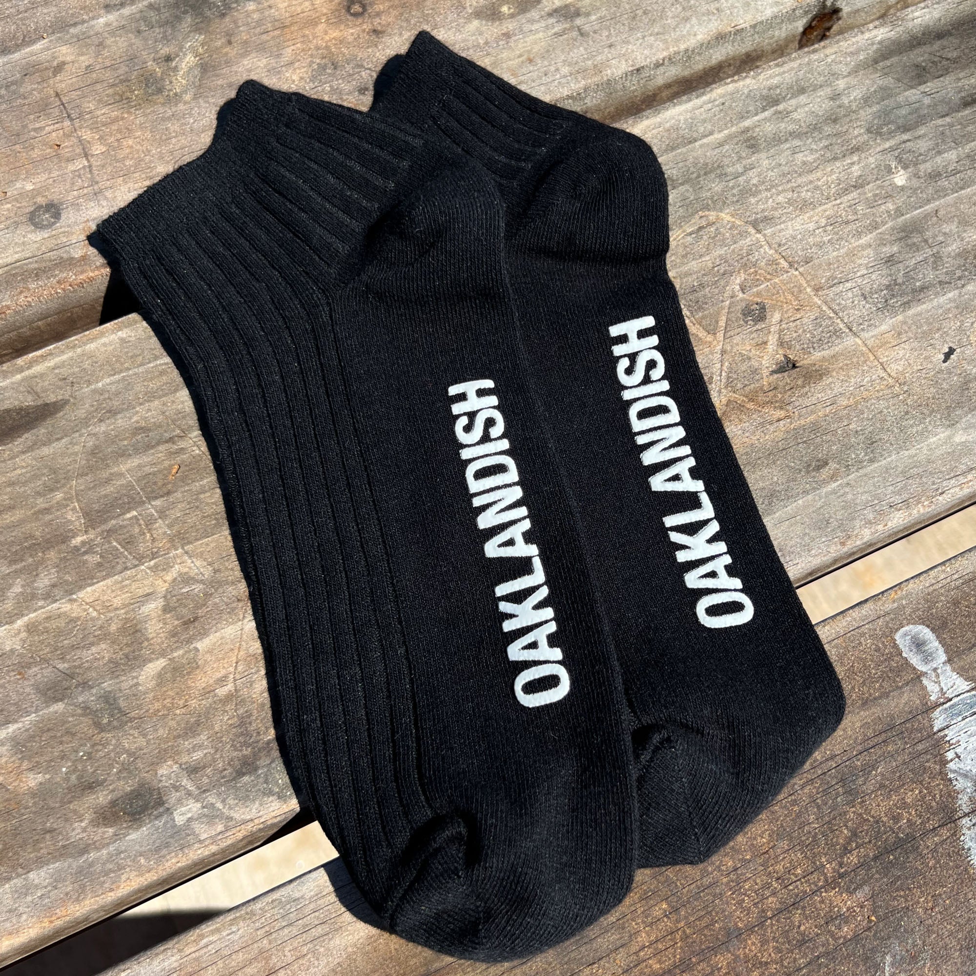 Men's Low Crew Socks in black without packaging. Oaklandish text across sole. Socks positioned on wooden bench in natural light.