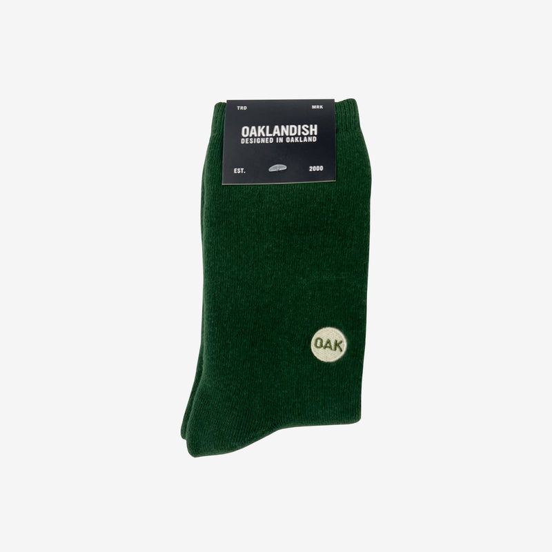 Plush high-cut green crew socks with an embroidered Oak wordmark detail on the ankle folded in Oaklandish retail packaging.