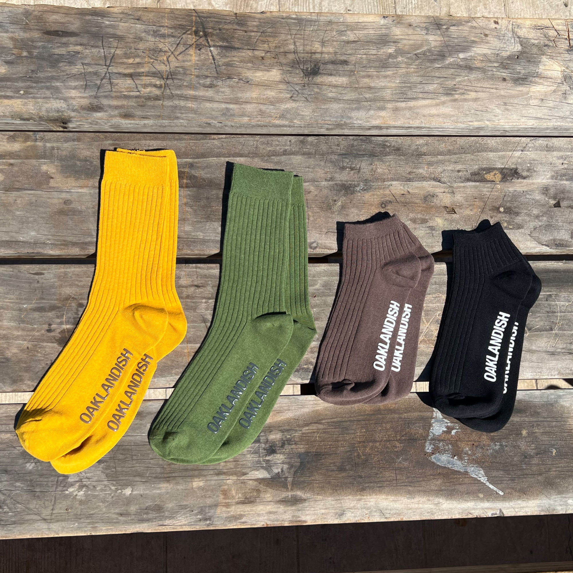 Men's Crew Socks in green, yellow, slate and black low grip sock included without packaging. Oaklandish text across sole. Socks positioned on wooden bench in natural light.