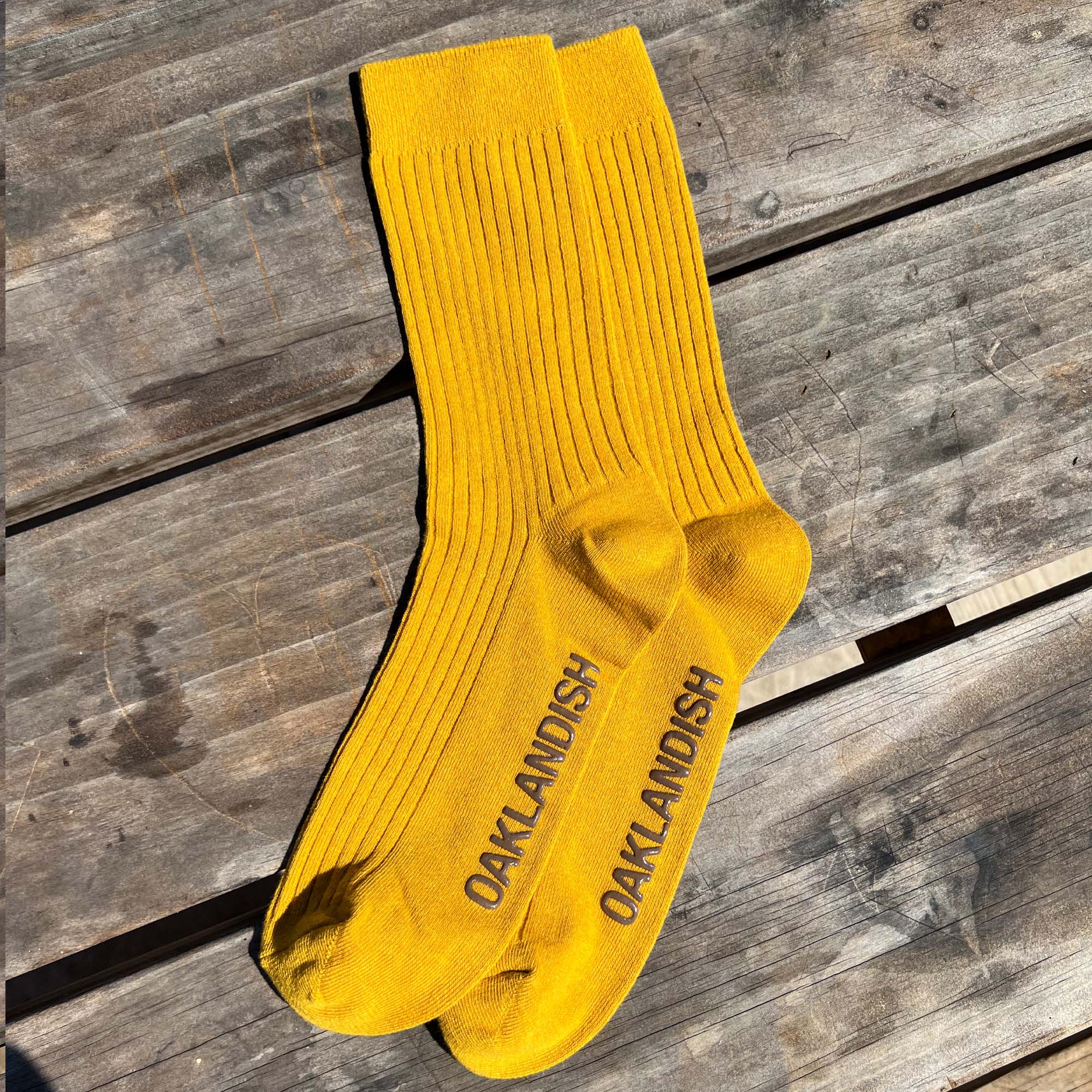 High cut yellow men's crew socks with Oaklandish wordmark in brown on sole. Socks positioned on wooden bench.