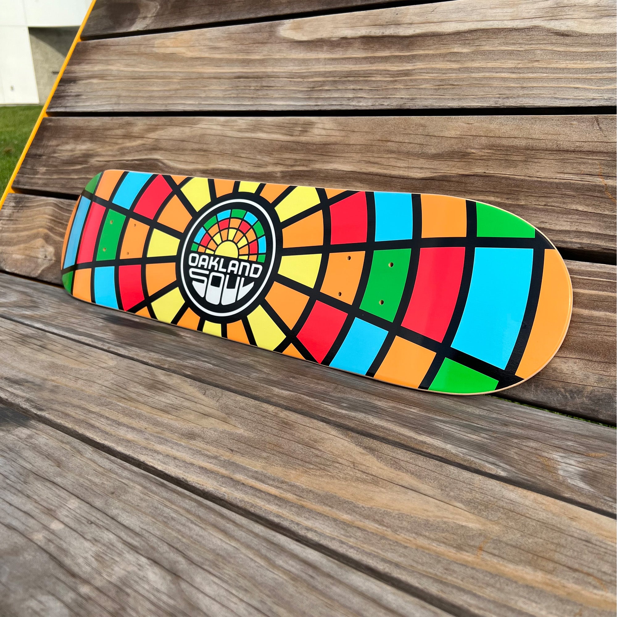 The Oakland Soul Logo Skate Deck rests on a wooden surface. Its 7-ply maple wood construction boasts a vibrant, stained-glass-like design with segments of orange, red, blue, and yellow. At the center, a circular logo reads "Oakland Soul SC," beautifully contrasting with the natural wood background.