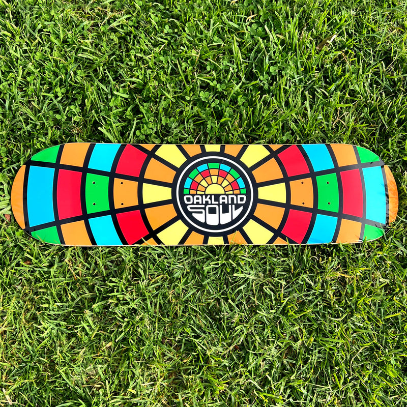 Outdoor photo image of skateboard deck with full-color Oakland Soul Soccer Club mosaic colors and round logo mark on grass.