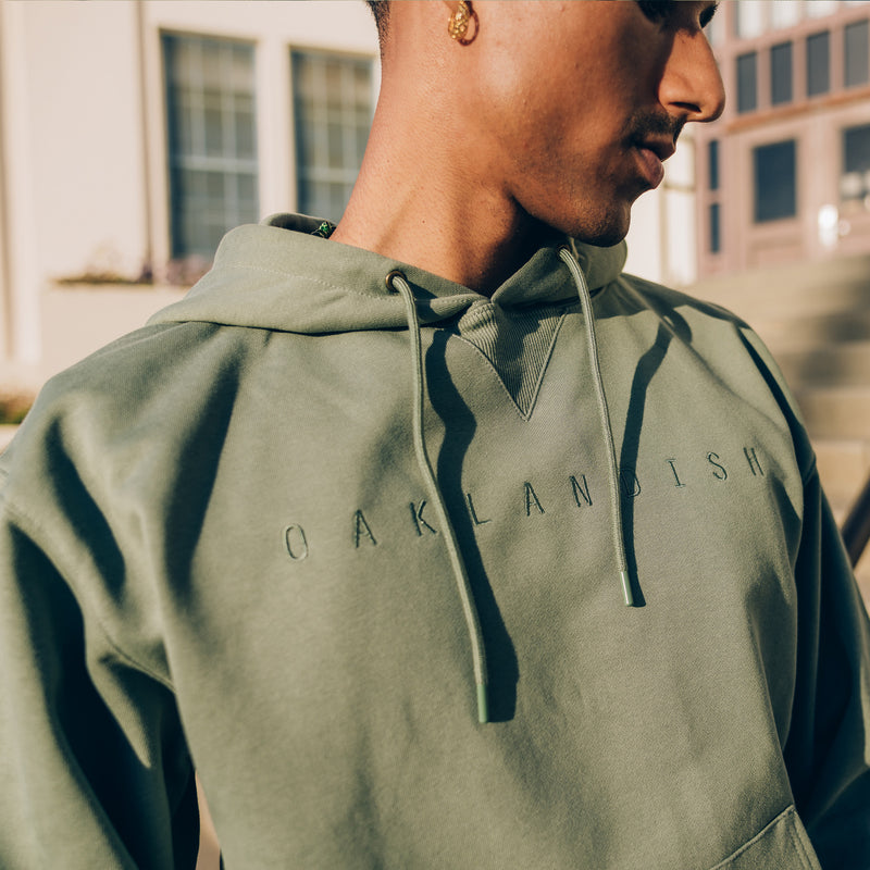 Detailed close-up of model wearing army standard pullover hoodie with monochromatic drawcords and Oaklandish embroidered across chest.