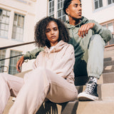 Man and woman sitting on outdoor steps wearing Oaklandish hoodie and jogger sets, with the woman in sand and man in army green.