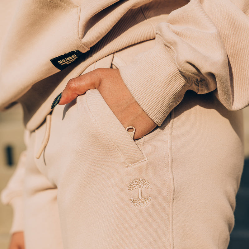 Detail close up of of sand standard sweatpants with model's hand in pocket of front zippered left pocket and embroidered tree logo.