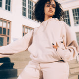 Woman standing outside in a sand-colored Oaklandish hoodie and jogger set. Hand in kangaroo hoodie pocket.