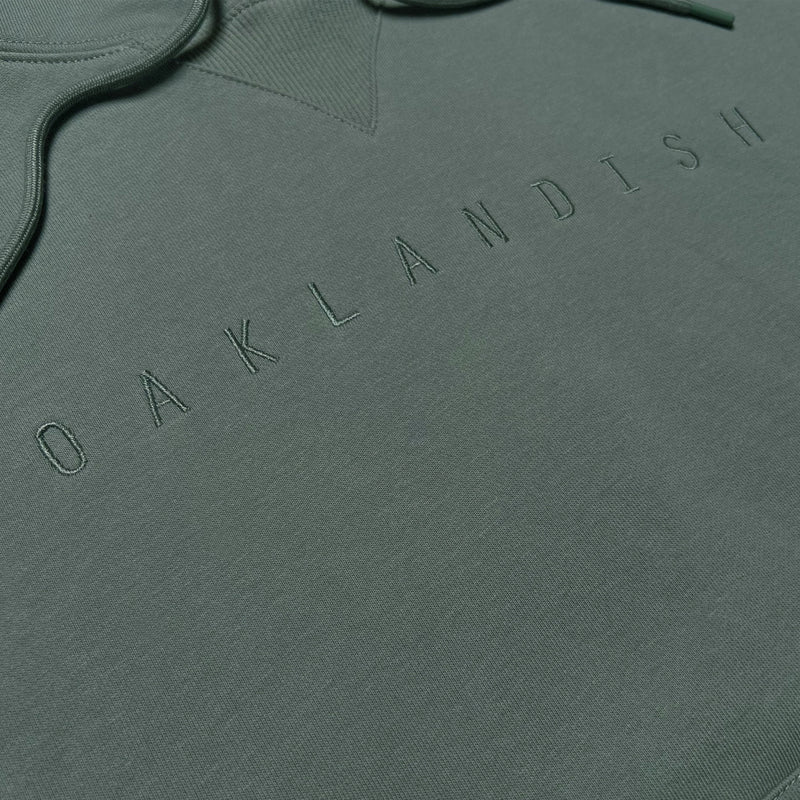 Detailed close-up of embroidered Oaklandish wordmark on an army green hoodie worn by a man.