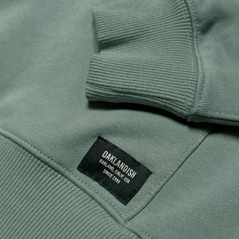 Detail close up of Oaklandish label on bottom of kangaroo pocket on Army green hoodie.