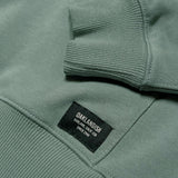 Detailed close-up of Oaklandish label on the bottom of kangaroo pocket and of the arm cuff ribbing on an army green hoodie.