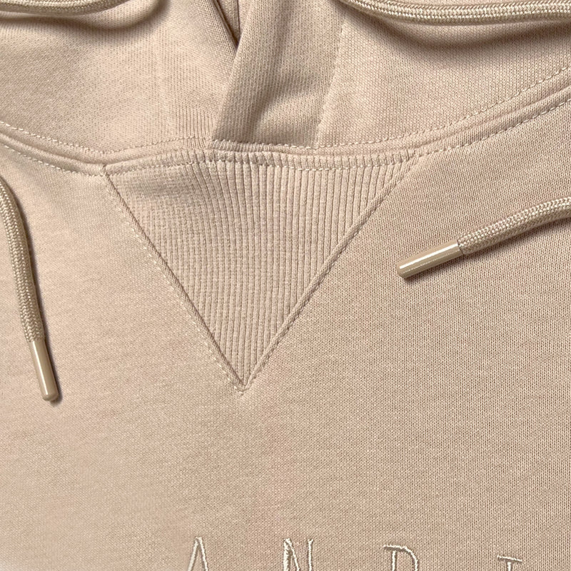 Detailed close-up of the triangle ribbing patch at the collar and metal drawcord tips on a sand-colored hoodie.
