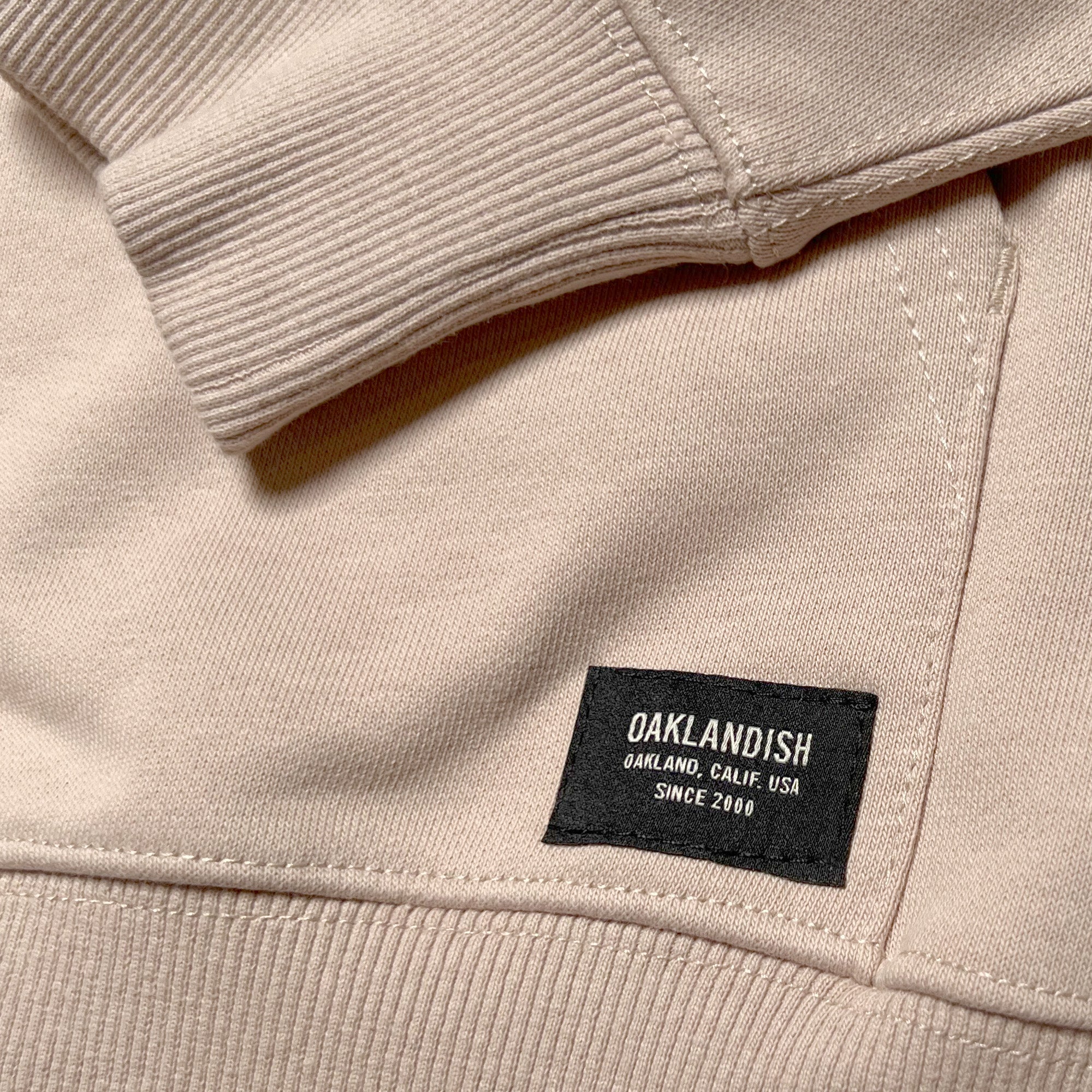 Detailed close-up of the triangle ribbing patch at the collar and metal drawcord tips on a sand-colored hoodie.