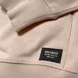Detailed close up of woven label at bottom left corner of kangaroo pocket and ribbing at cuff and waistband of sand pullover sweatshirt.