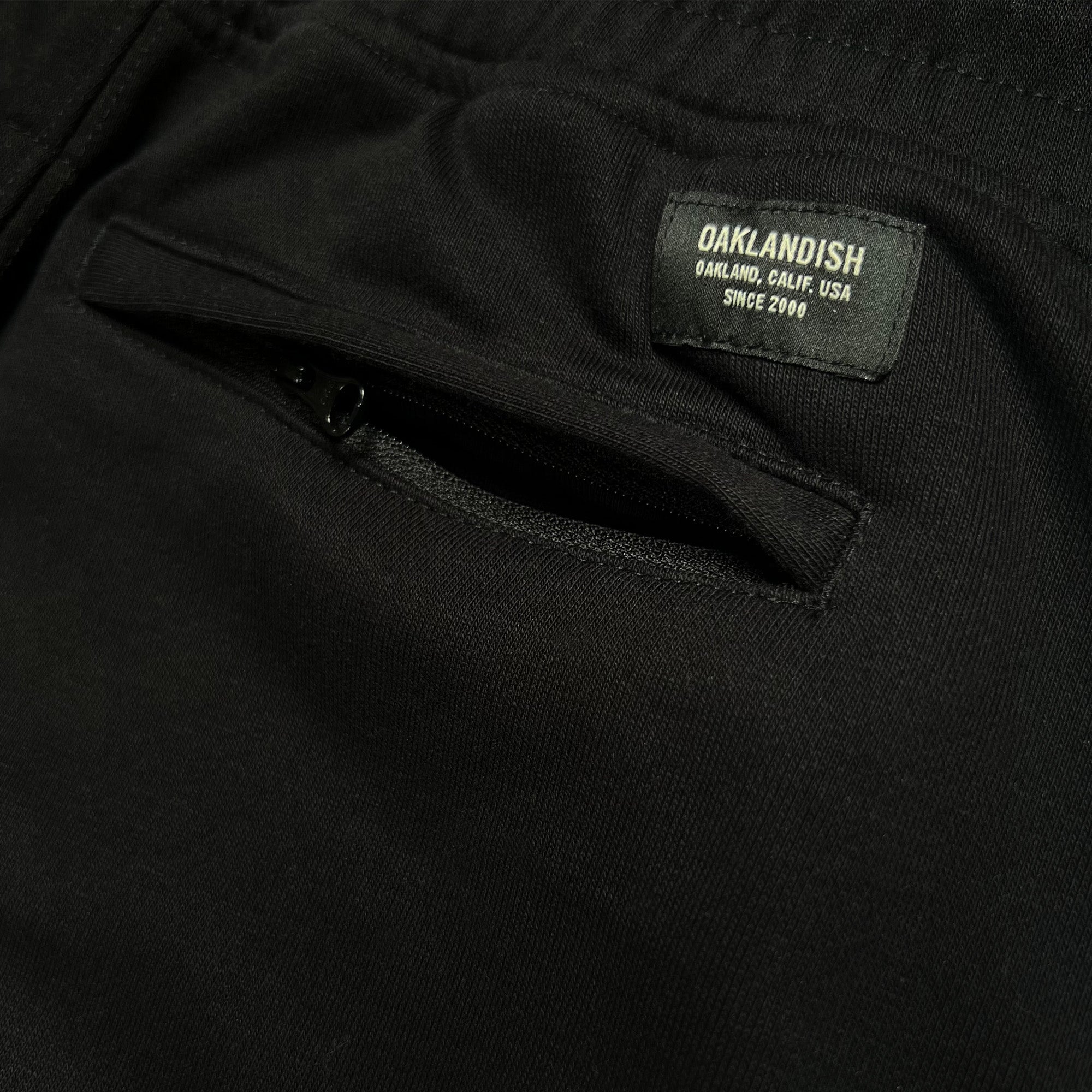 Detail close-up of the Oaklandish label above the back zipper pocket of black joggers.