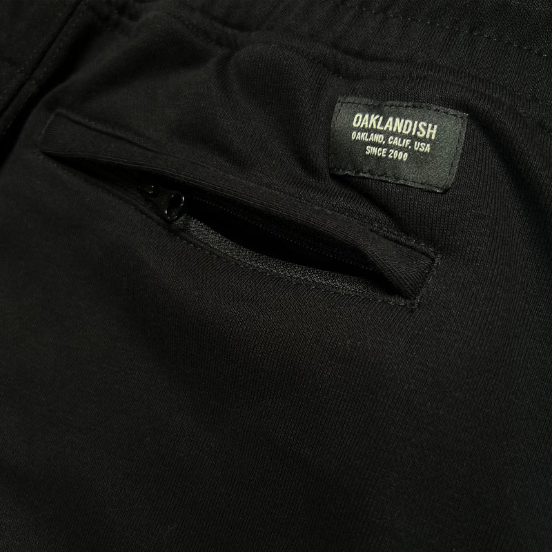 Close-up of Oaklandish label on the backside of black joggers with zippered back pockets.